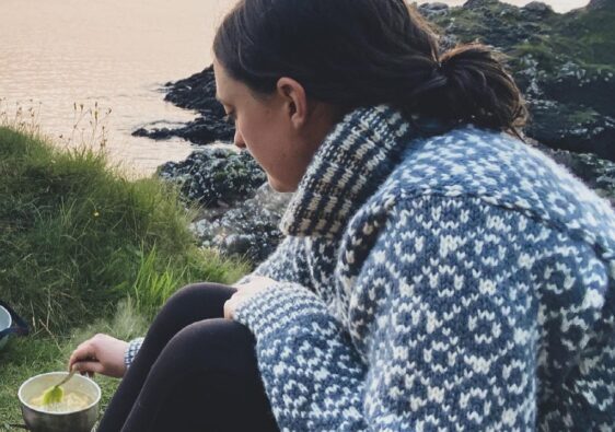 Heather sitting on a cliff near Oban in Scotland wearing a big wooly jumper and cooking over an outdoor stove.