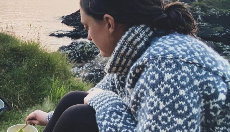 Heather sitting on a cliff near Oban in Scotland wearing a big wooly jumper and cooking over an outdoor stove.