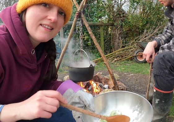 Heather is holding a stainless steel bowl of wild garlic dumplings in front of a campfire. She is wearing yellow hat and a maroon hoodie and is smiling.